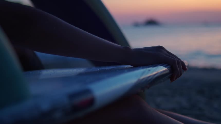 Hand holding a surfboard by the sea during sunset