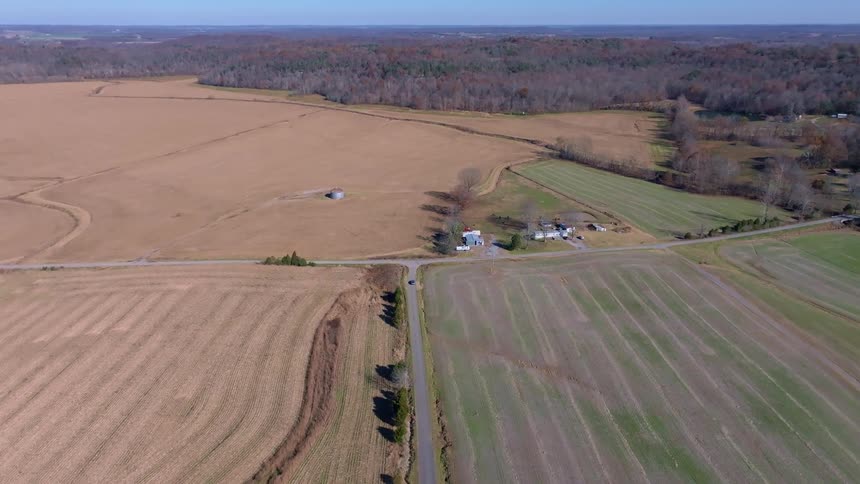 An aerial drone shot flies over a flat rural landscape with sections of green and brown fields a road runs through the center of the frame with a few houses and buildings visible among the fields and patches of trees