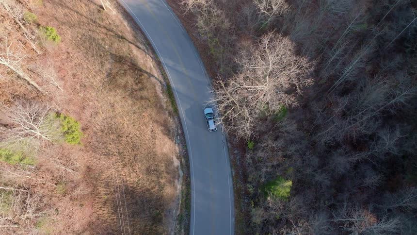 The video shows a high aerial shot of a darkcolored car driving on a winding paved road the road is surrounded by fields and trees with autumn foliage the video also has a watermark that reads filmsupply