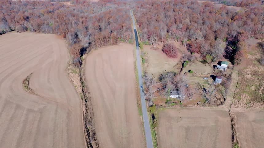 Narrow road cuts through fields and an area of autumncolored trees