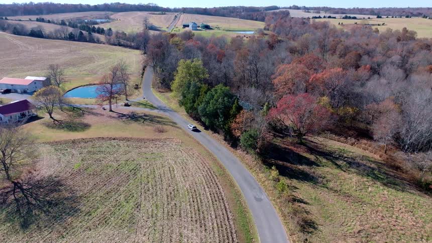 Rural road runs between houses a pond and a wooded area with autumn colors