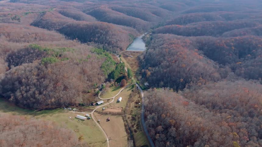 An aerial view captures rolling forested hills with autumn foliage a winding road scattered farm buildings and a small lake nestled in the distance