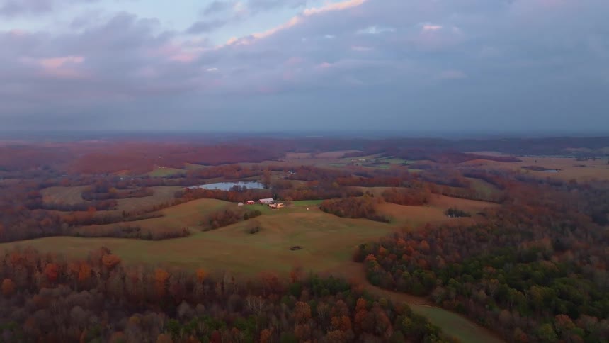 An aerial shot captures a rural landscape with rolling hills and patches of deciduous trees in autumn colors a small cluster of buildings is visible in a clearing with a pond nearby under a dramatic cloudy sky