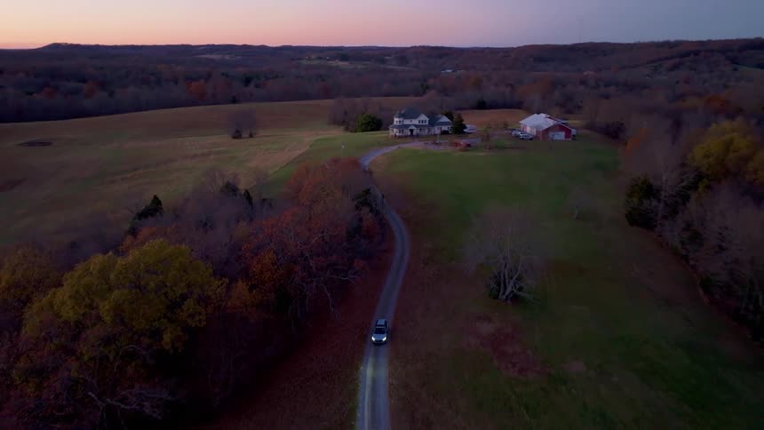 The video shows a high aerial shot of a white car driving down a dirt road the road winds through a hilly forested landscape the sun is setting and there is a large house and a red barn visible in the distance the video has a watermark that reads f