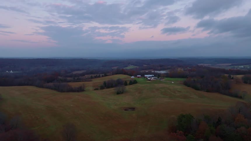 The video is a highangle shot of a rural landscape at dusk featuring rolling hills and forested areas a cluster of buildings including what appears to be a farmhouse with a red roof is visible on top of one of the hills the sky is a mix of muted pin