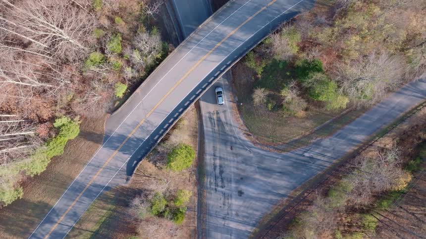 An aerial drone shot follows a car as it drives along a narrow winding road that goes underneath a larger overpass the surrounding landscape is a mix of bare trees and some evergreens suggesting the autumn or winter season