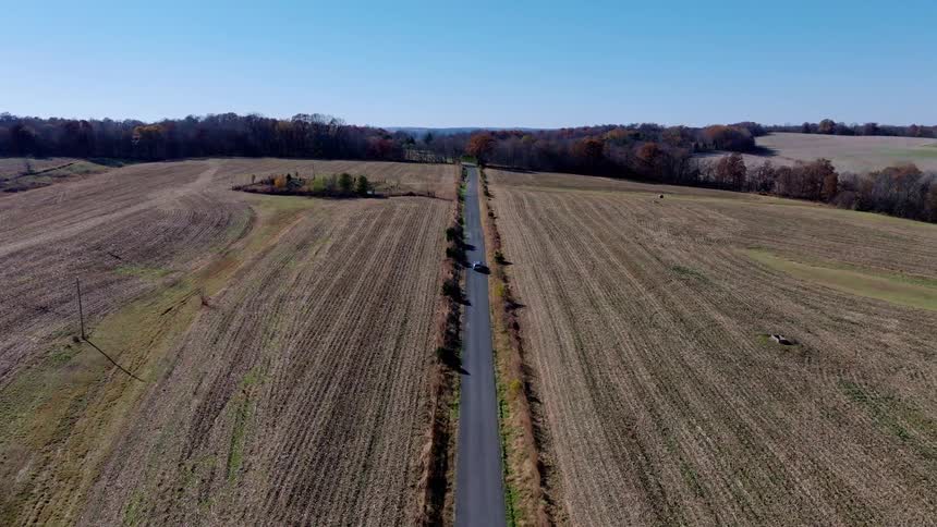 An aerial shot follows a car as it drives down a long straight road through a rural landscape the road is flanked on both sides by fallow fields and bare trees with a small pond visible in the distance