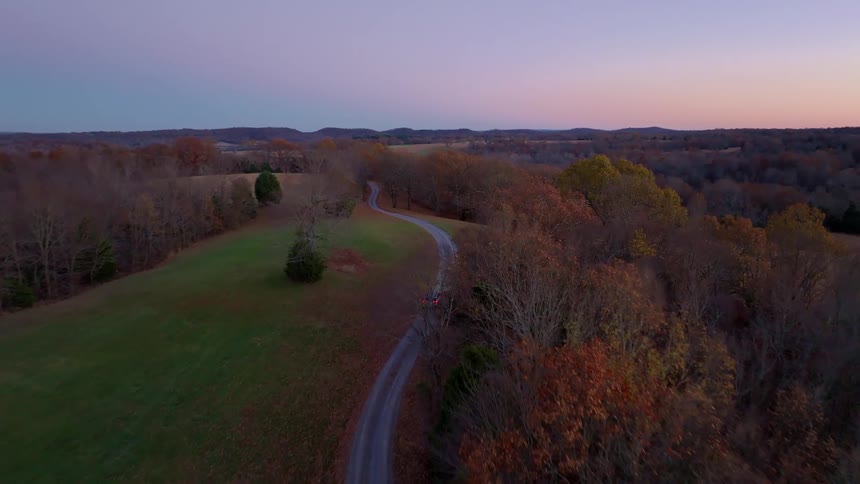 In the evening a car drives down a lonely road in a rural setting lined with trees fallen leaves and grasses
