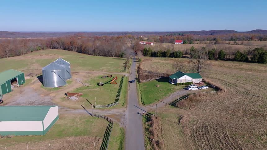 An aerial view of a car driving on a paved road in a rural area with a farm with silos on the left side