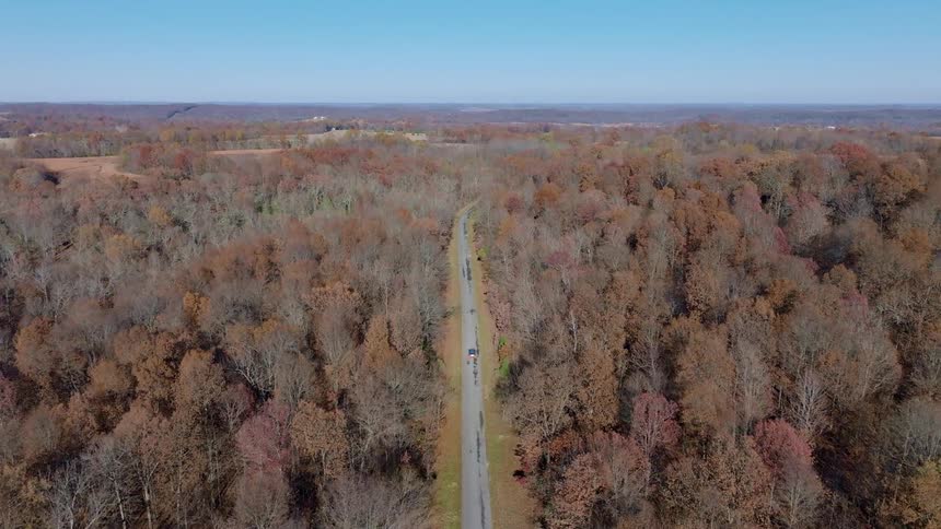 The video shows a high aerial shot of a long paved road surrounded by a forest of leafless trees a darkcolored suv is driving in the distance
