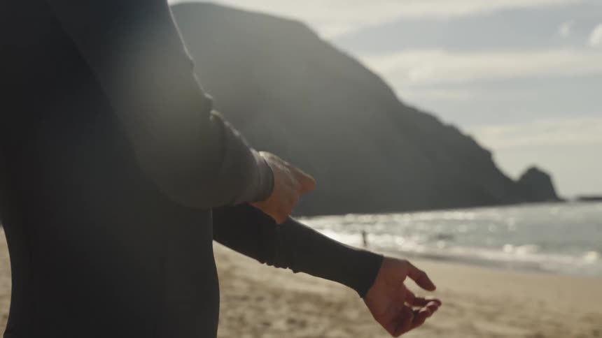 A shot of a person on a beach shows them pulling up the sleeves of a wetsuit the background features a sandy shore the ocean and a large hill or cliff face under a bright sky