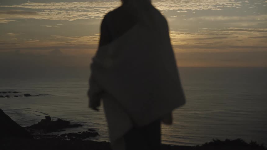A young woman walks down a rocky coastline at sunset