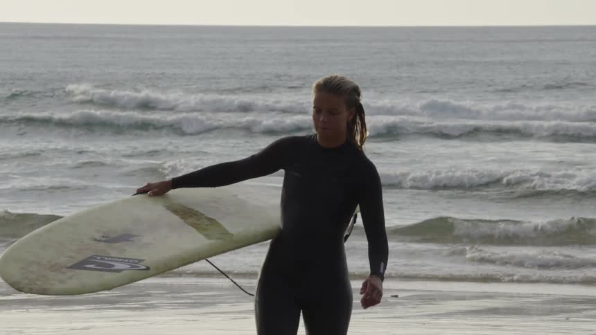 A female surfer walks out of the ocean onto the beach wearing a wetsuit and holding a surfboard smiling at midday