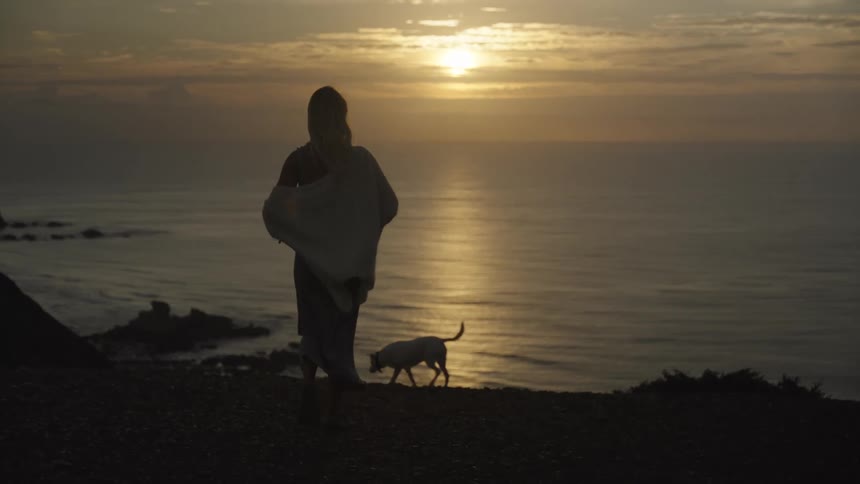 Woman walks along the coast with her dog as the setting sun shines through clouds