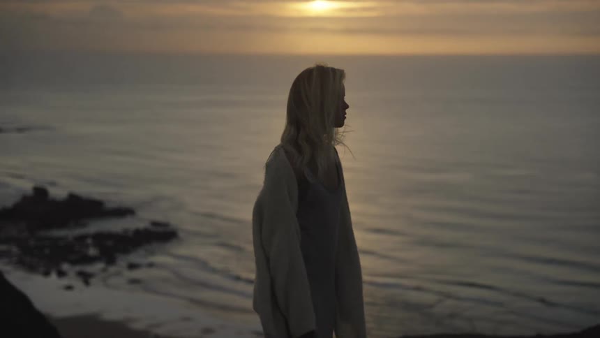 A woman near the edge of a cliff above a bay beach looks out to the sunset then turns around