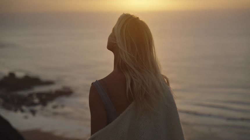 A woman with her hair blowing in the wind standing on a rock in the ocean at sunset