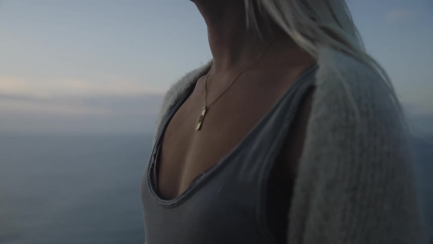 A woman in a gray top and light sweater stands by the ocean at dusk with the wind blowing her hair