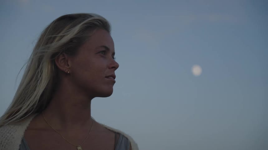 A blonde woman stands outdoors at sunset with a light wind blowing through her hair