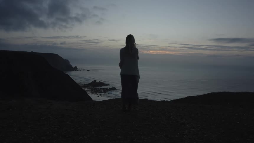 Woman standing on a cliff watches the sea at dusk