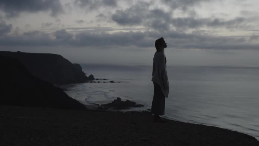 Woman standing along the coast looking up at the cloudy sky