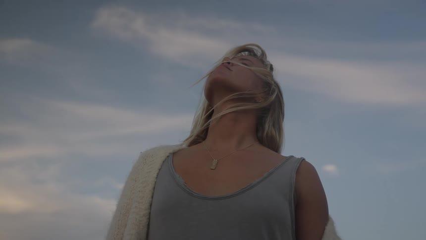 A woman in a gray top and light sweater tilts her head upward toward the sky with her hair blowing in the wind