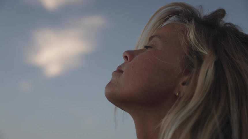 A woman with blonde hair tilts her head back with closed eyes peacefully facing the sky