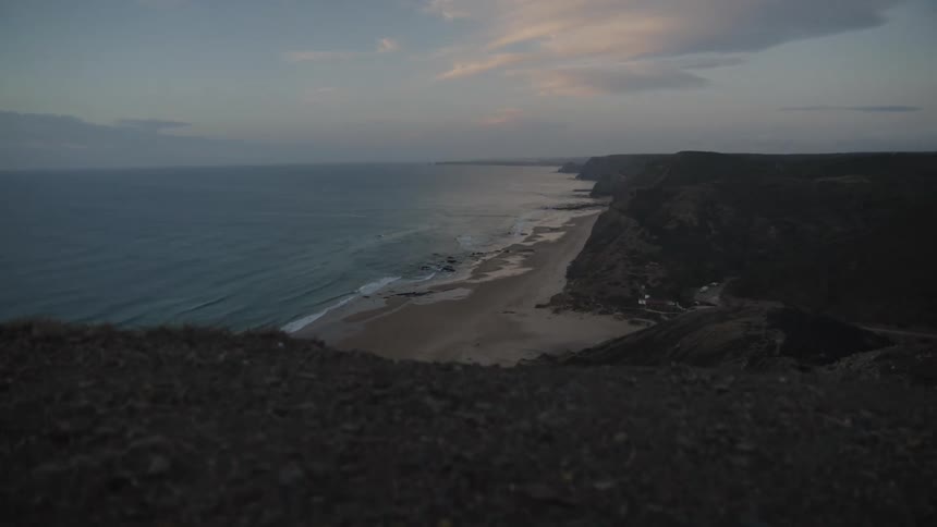 The video shows an aerial view of a dramatic coastline at sunset the camera is looking down at a large cliffside and the ocean below