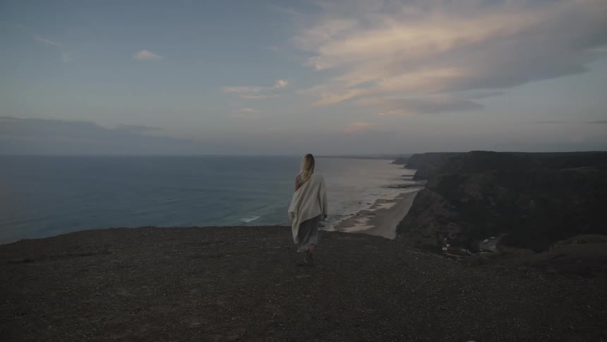 A woman draped in a sweater walks with a white dog along a cliffside overlooking the ocean at sunset