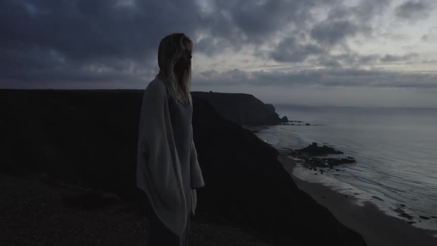 A blonde woman stands on a cliff overlooking the ocean at sunset