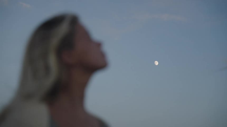 A blonde woman initially out of focus who comes into sharp focus as she looks up and inhales deeply under a serene dusk sky with a visible moon