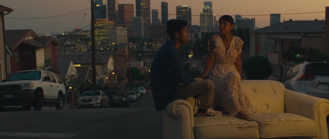 A young couple sitting on a couch placed on a street in a suburban area conversing against the backdrop of a distant litup city skyline at dusk