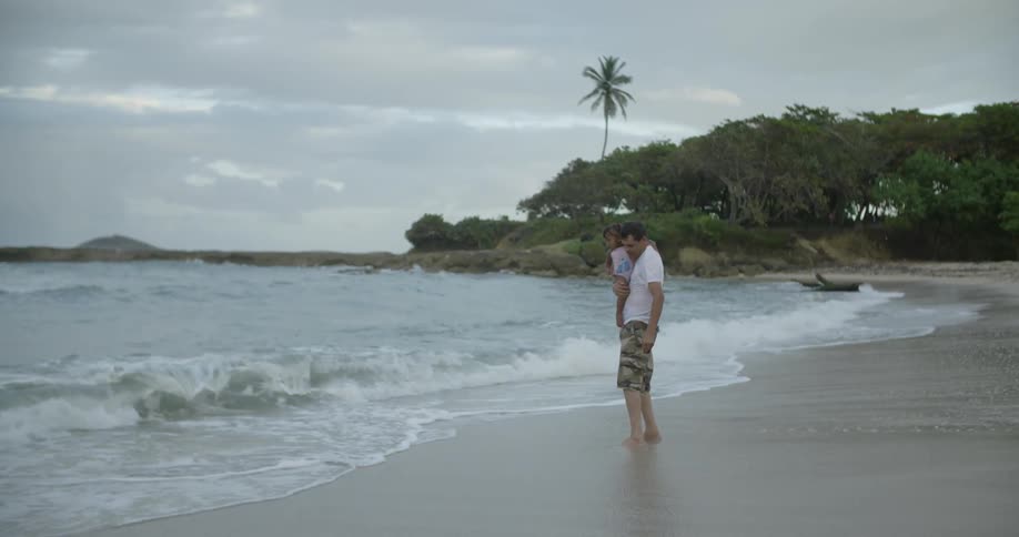 A man holds a baby on the beach as waves come ashore