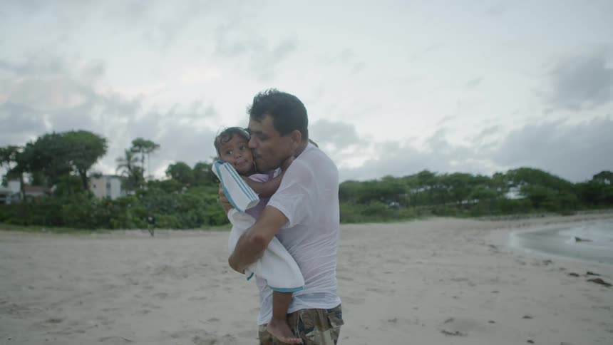 A man is standing on a beaching holding and kissing his daughter