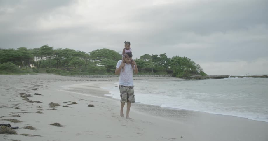 Man carrying toddler girl on shoulders on beach on cloudy day
