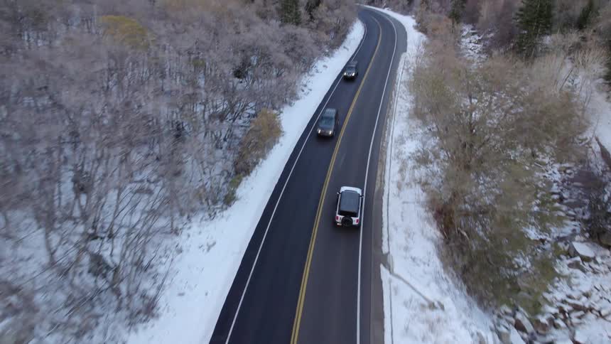 An aerial view of a white jeep driving on an asphalt road with forests on the sides covered in snow on the ground