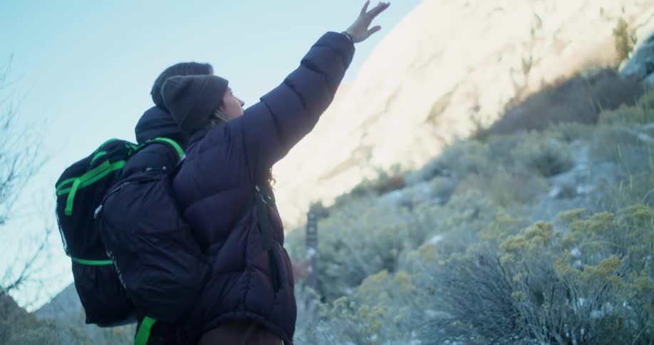 Two individuals with backpacks are standing on a hill in a rugged mountainous area they are looking up and gesturing toward the rocky cliff face in front of them