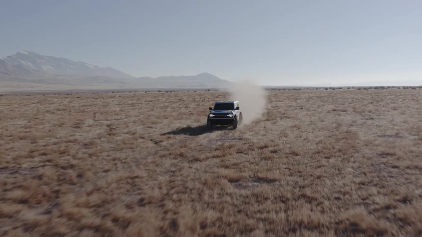 White suv driving along a dirt trail in a barren field with mountains in the background