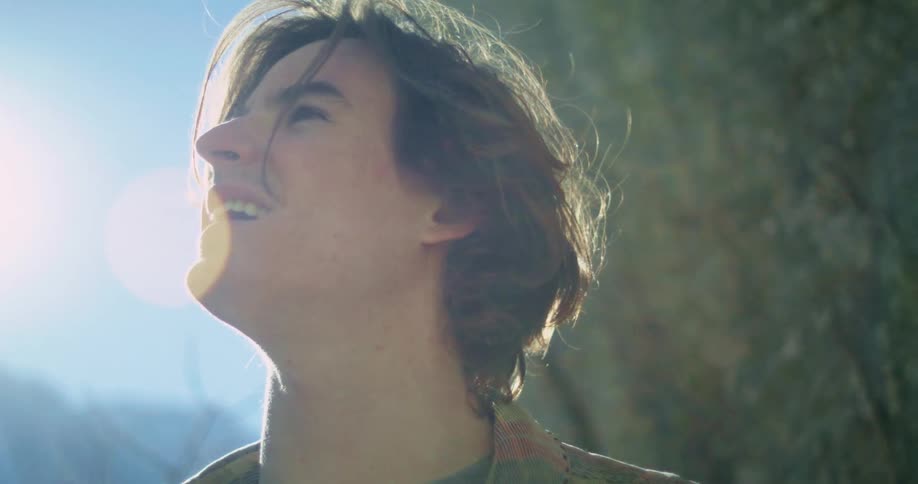 The video shows a person with long brown hair and a plaid shirt looking up and smiling into the bright sun which creates a prominent lens flare the person then looks down the scene is outdoors with a rocky texture visible in the background the wate