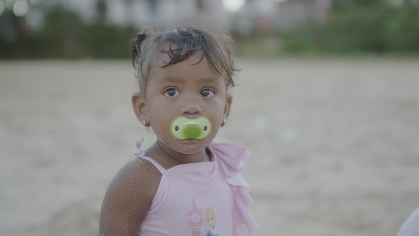 A toddler girl is standing on a beach and sucking on a pacifier