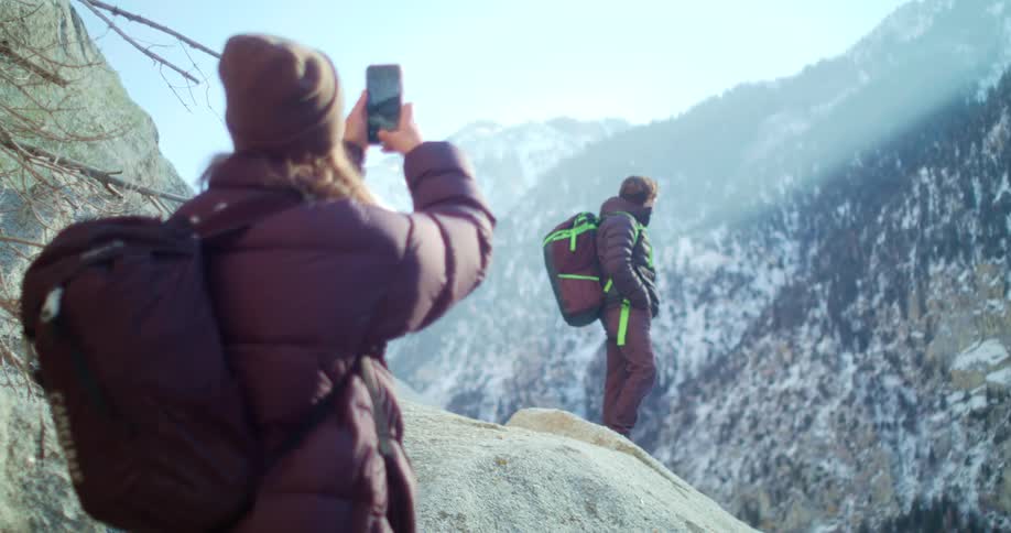 The video shows a person from behind wearing a beanie and a brown puffer jacket with a backpack holding a phone to take a photo in the foreground another person with a greenandblack backpack is posing for the photo on a rock