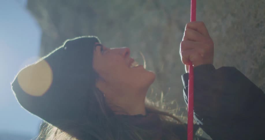 The video shows a closeup of a person wearing a dark beanie and jacket looking up with a hopeful expression they are holding onto a red rope which is taut the background is a rocky cliff face and there is a bright lens flare from the sun which is b