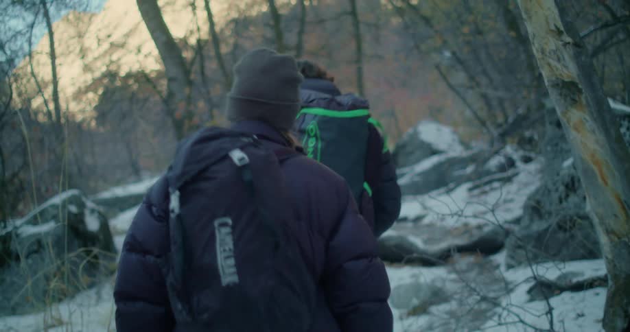 Two people are hiking in a snowy wooded area one person is wearing a dark puffer jacket with a camelbak backpack and a beanie