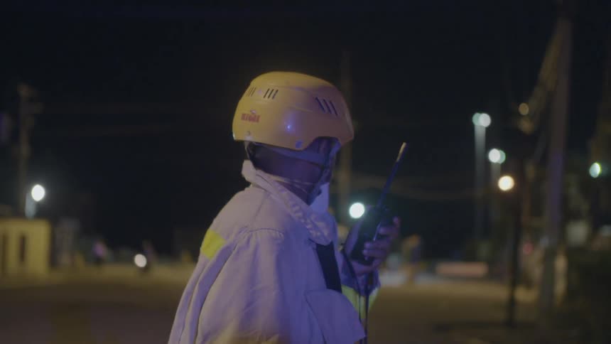 A firefighter stands outdoors with a walkie talkie with lights flashing around him