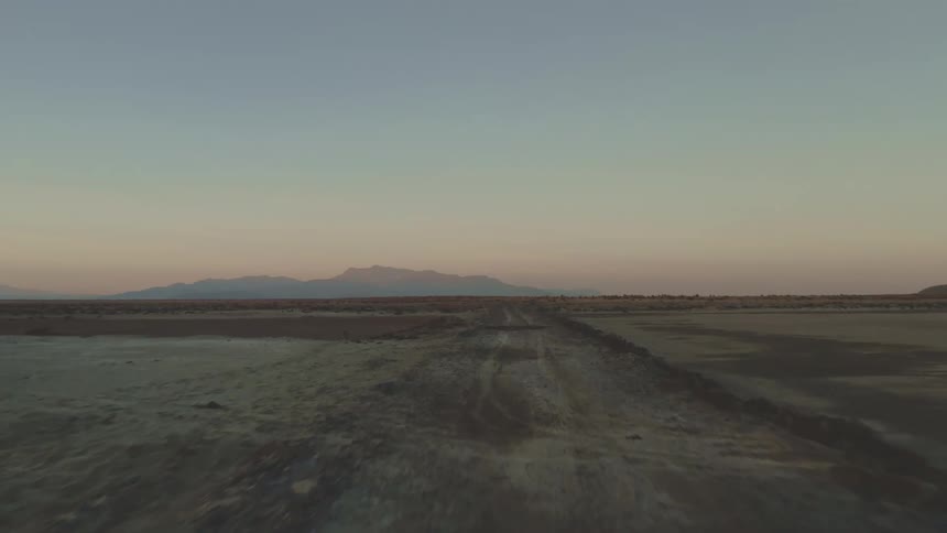 A barren dirt road stretches toward distant mountains under a soft evening sky