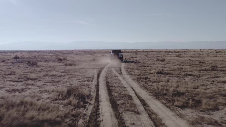 A white suv drives across dusty desert tracks toward distant mountains under a pale sky