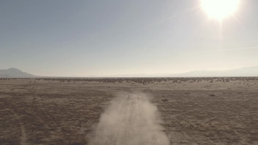 The video shows an aerial view of a black suv driving on a dirt road through a barren landscape the sun is bright in the background and the sky is hazy