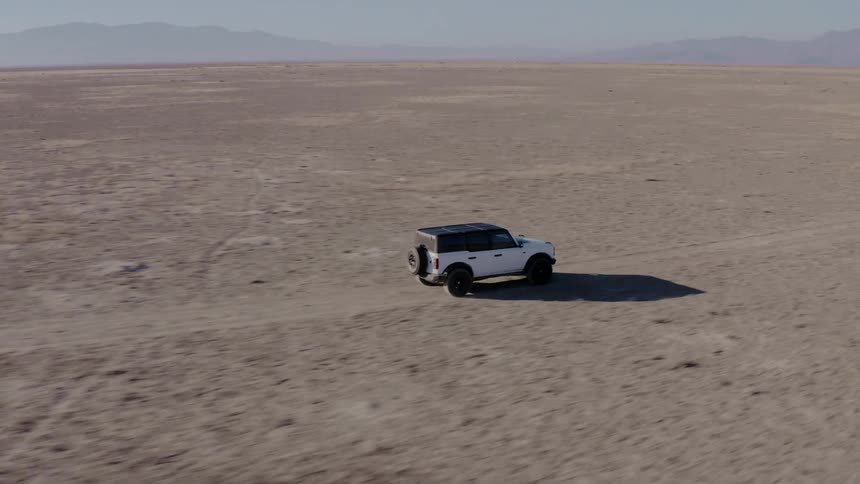 An aerial shot follows a white ford bronco as it drives across a wide flat and arid landscape mountains are visible in the distance under a clear sky