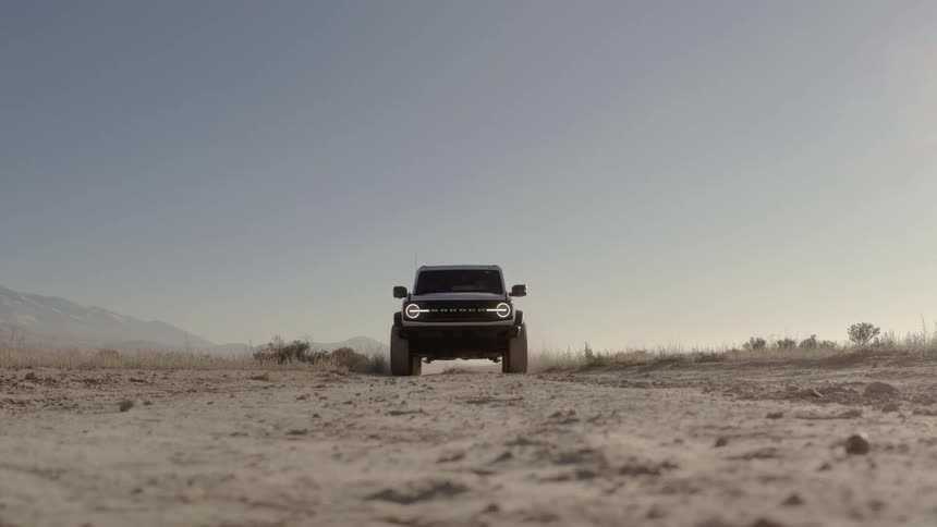 A black ford bronco driving toward the camera on a dusty dirt road in a vast arid landscape under a hazy sky