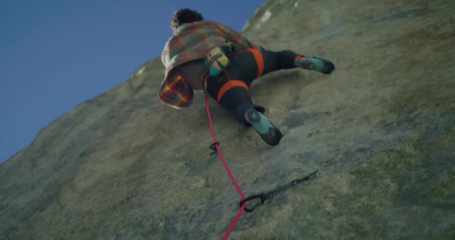 A man wearing a harness and climbing shoes ascends a steep rock face with a safety rope clipped into anchors