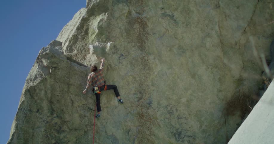 A male rock climber is scaling a large rock face outdoors he is wearing a plaid shirt and is attached to a red safety rope